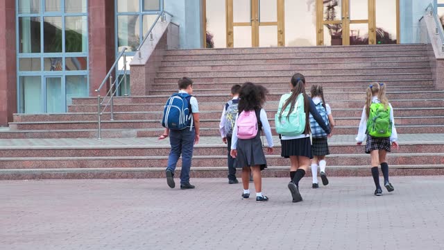 junior students in uniform with schoolbags walk to school building with stone steps on warm day backside view slow motion